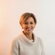 Close-up portrait of a smiling German woman in her late 30s, wearing a light sweater, with a soft peach and white background.